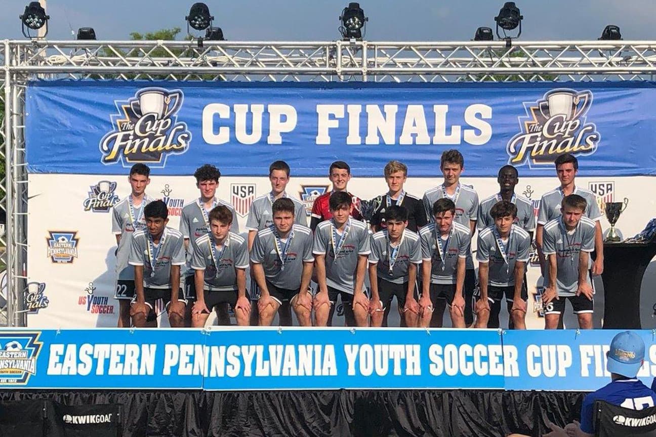 A youth boys' soccer team posing on a stage after the Eastern Pennsylvania Youth Soccer Cup Finals. The players are wearing grey jerseys and medals, standing in front of a blue and white "Cup Finals" banner.