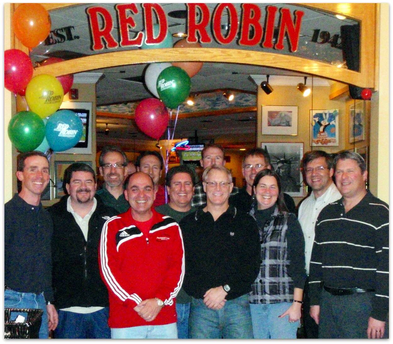 A group photo of twelve people standing in front of a Red Robin restaurant entrance. Chuck Nace is in the front center wearing a black shirt.