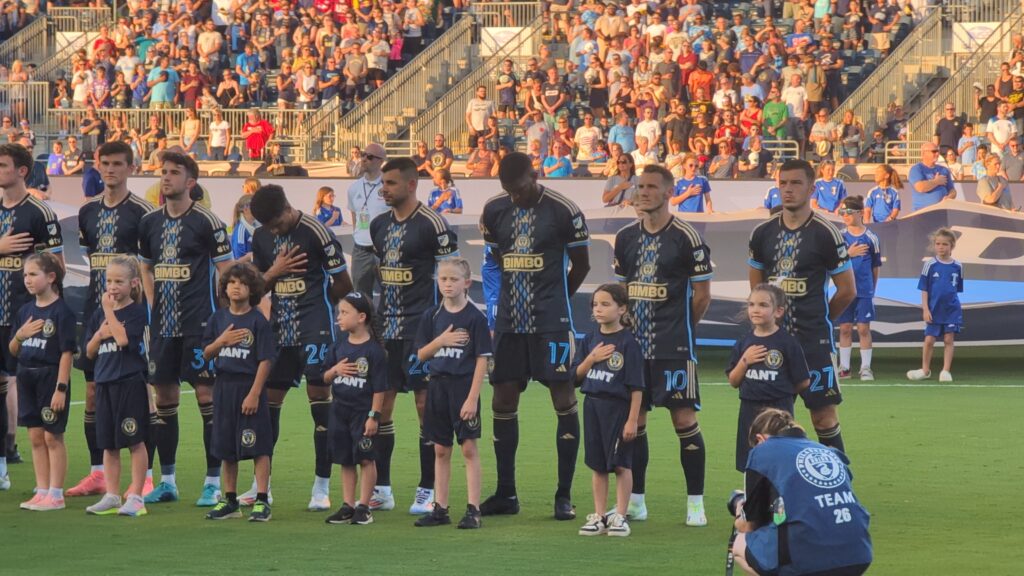 A row of Philadelphia Union soccer players and young children standing on a field with hands over their hearts for the national anthem. Fans fill the stadium bleachers in the background.
