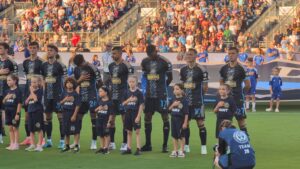 A row of Philadelphia Union soccer players and young children standing on a field with hands over their hearts for the national anthem. Fans fill the stadium bleachers in the background.
