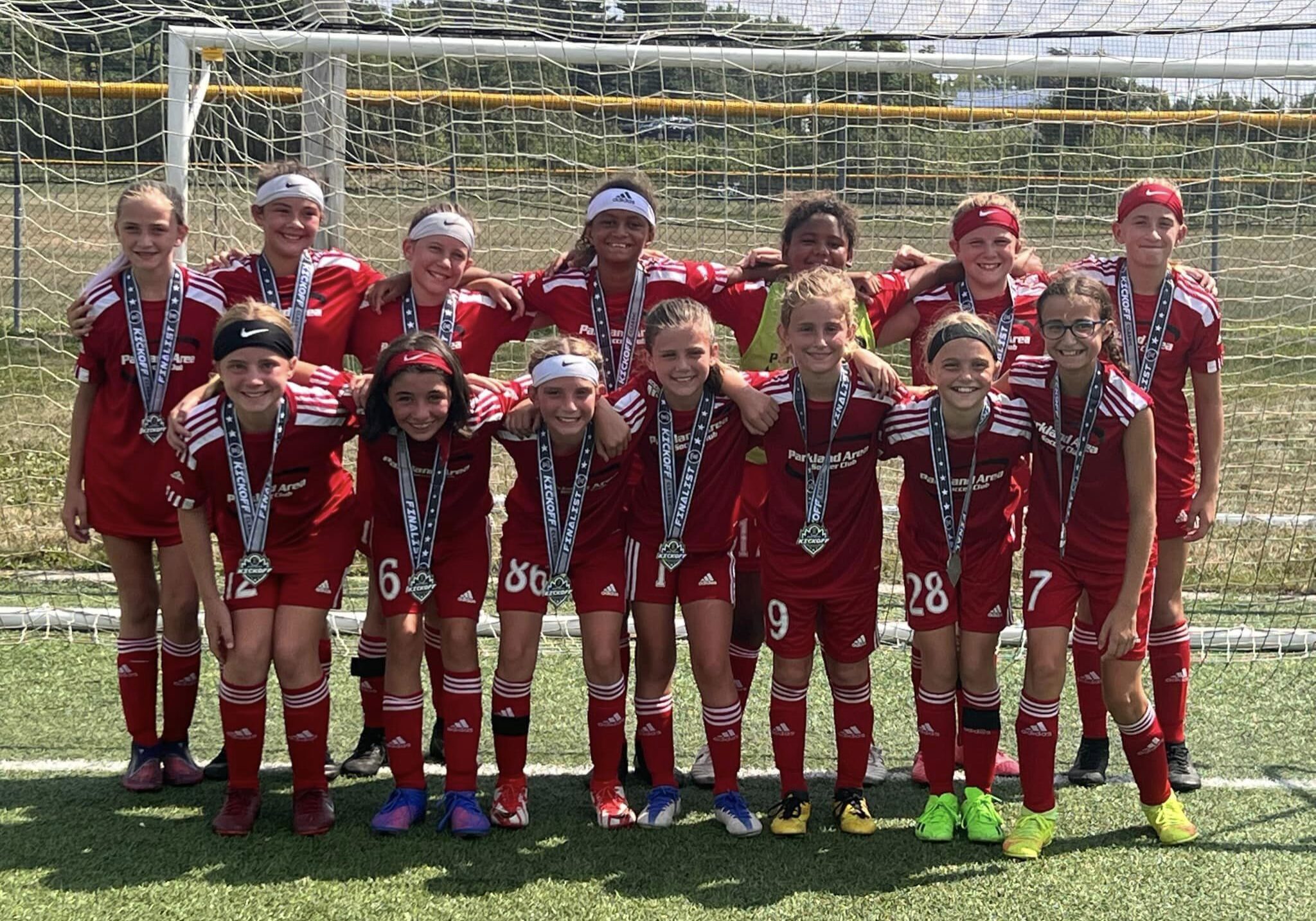 A group of young girls from the Parkland Area Soccer Club posing for a photo in front of a soccer goal. They are wearing red uniforms and silver medals after a tournament.